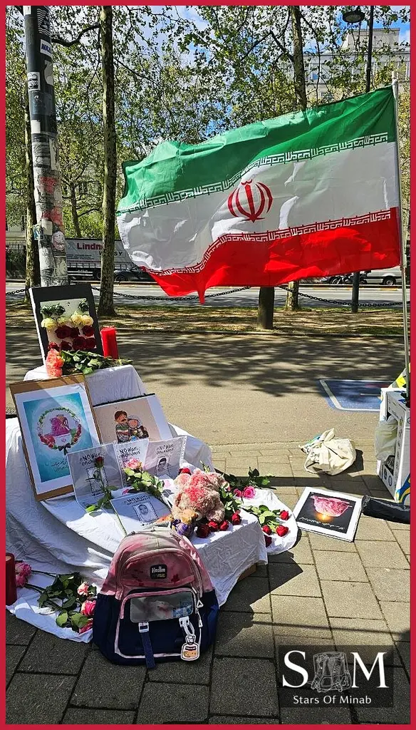 A poignant outdoor display at a Brussels Minab peace gathering, featuring a waving Iranian flag, a table laden with red and pink roses, lit candles, and framed drawings of children and families. A child