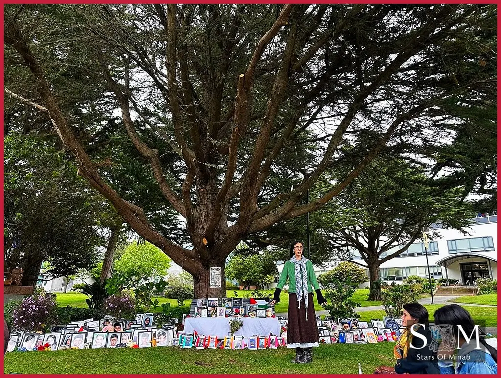 Wide view of a tree-shaded campus memorial with photos and candles at the San Francisco State University vigil, a student stands in reflection among tributes